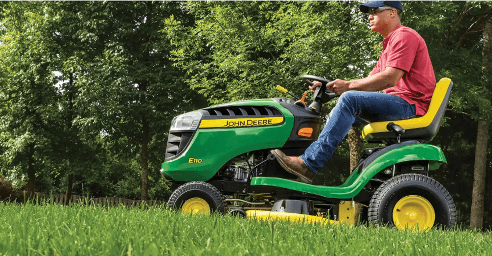 Man riding a green John Deere lawn mower on a grassy lawn, surrounded by trees.