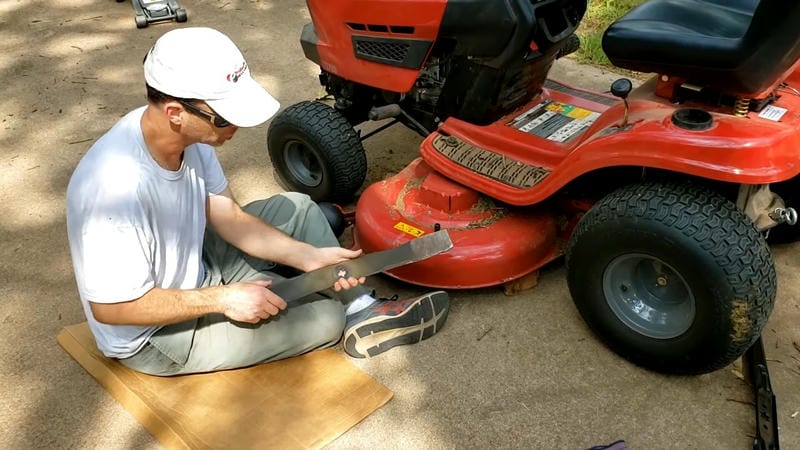 Man sharpening a lawn mower blade while sitting next to a red riding mower on a sunny day.