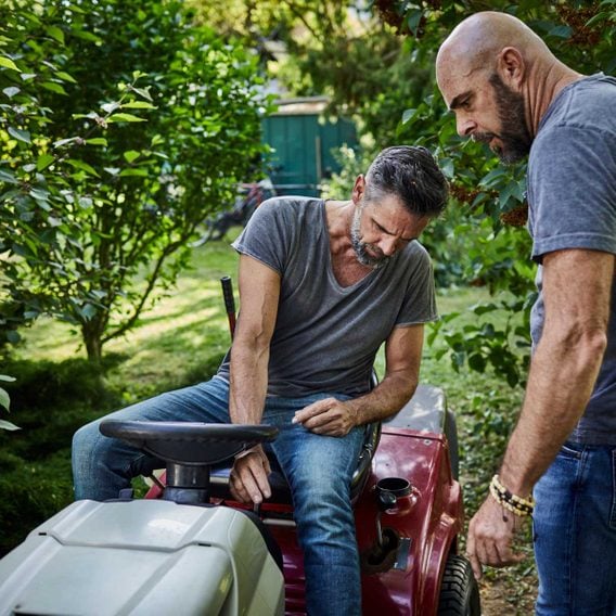 Two men working together on a riding lawn mower in a lush garden, with one seated and the other standing.