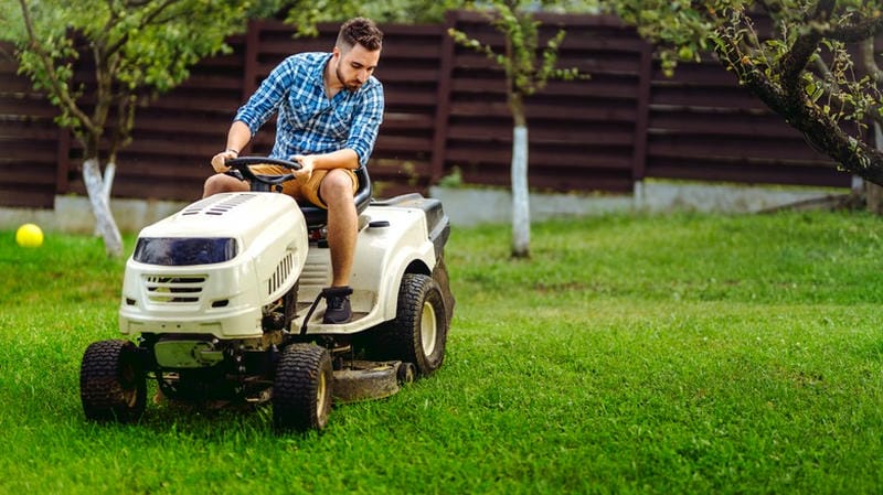 Man driving a white riding lawn mower to cut grass in a backyard with trees.