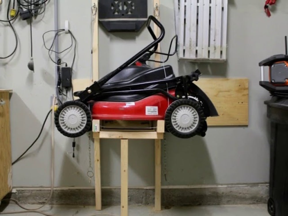 Red lawn mower stored vertically on a wooden stand against a garage wall.