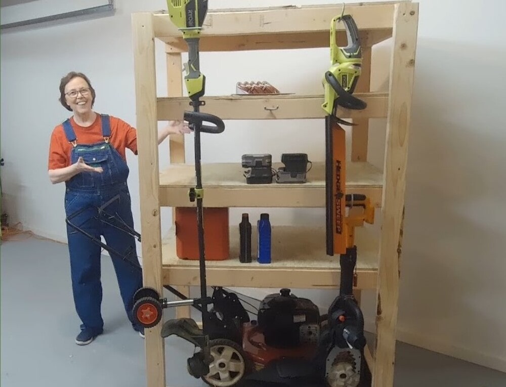 Smiling woman standing beside a wooden storage shelf holding various garden tools, including a lawn mower and trimmers.