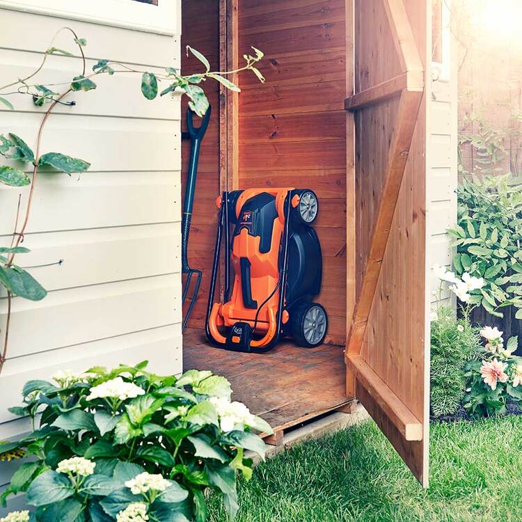 Orange lawn mower stored vertically inside a small wooden shed with garden tools.
