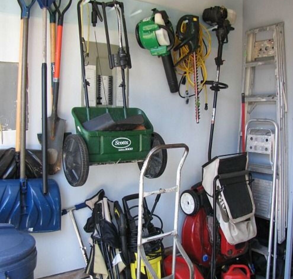 Neatly organized garage corner with garden tools, ladders, and equipment, including a green spreader.