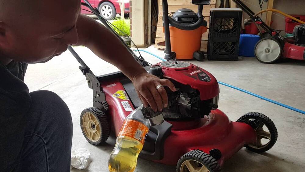Man draining fuel from a red lawn mower into a plastic bottle in a garage