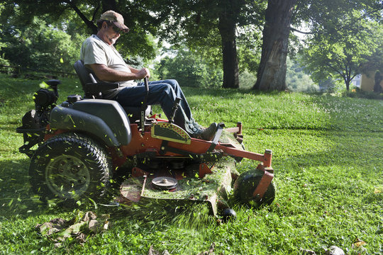 Man riding a zero-turn lawn mower while cutting grass in a shaded, tree-filled yard.
