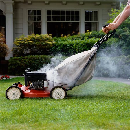 Lawn mower releasing smoke while cutting grass in front of a house, indicating a mechanical issue