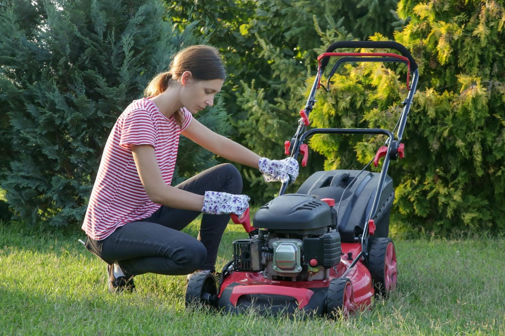 Woman checking the engine of a red lawn mower in a garden, preparing for maintenance.