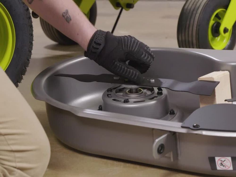 Gloved person installing a lawnmower blade onto its housing on a workshop floor.