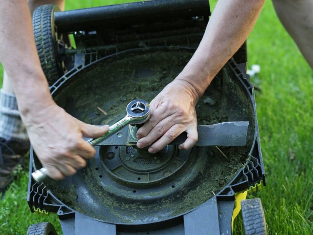 Person using a wrench to remove a lawnmower blade for maintenance on grass.