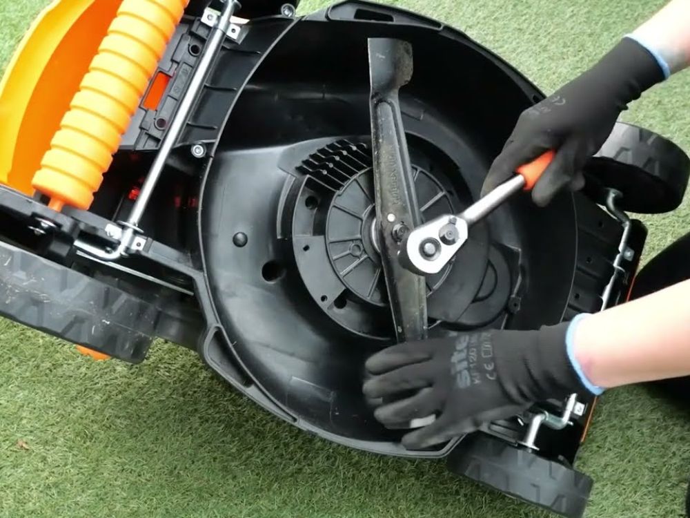 Gloved person removing a lawnmower blade with a wrench on artificial grass