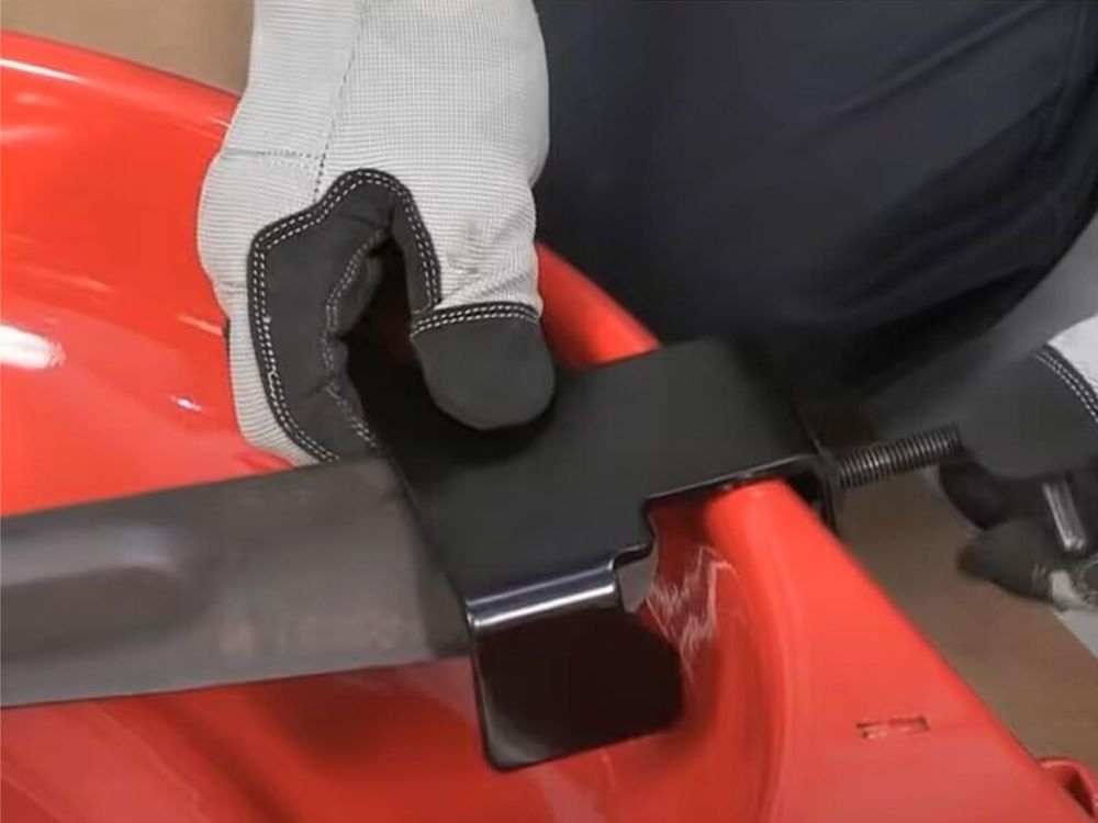 Gloved person using a blade removal tool to secure a lawnmower blade on a red deck.