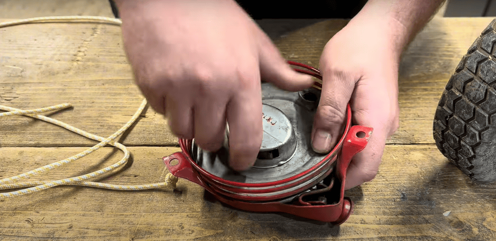 Hands adjusting the pull cord mechanism of a lawn mower on a wooden workbench