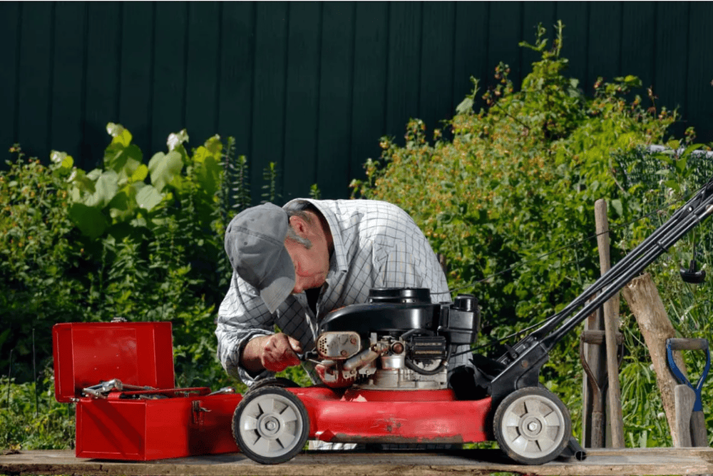 Older man examines a lawn mower engine, surrounded by tools and greenery