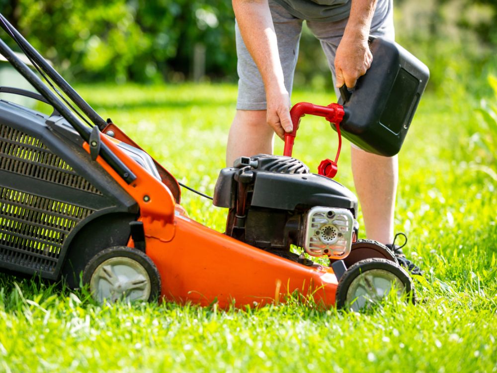 Person refilling an orange lawn mower with fuel using a black gas can on a green lawn.