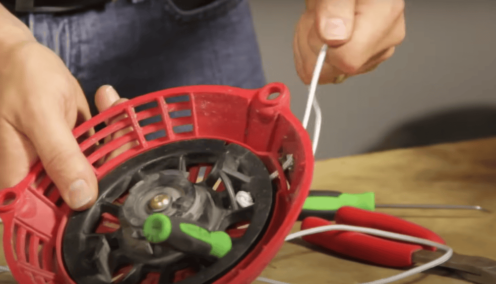 Hands threading a cord through a lawn mower recoil starter, surrounded by tools